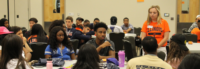 A man holds a microphone at a circular table while a room of college-aged indivuals look on. 